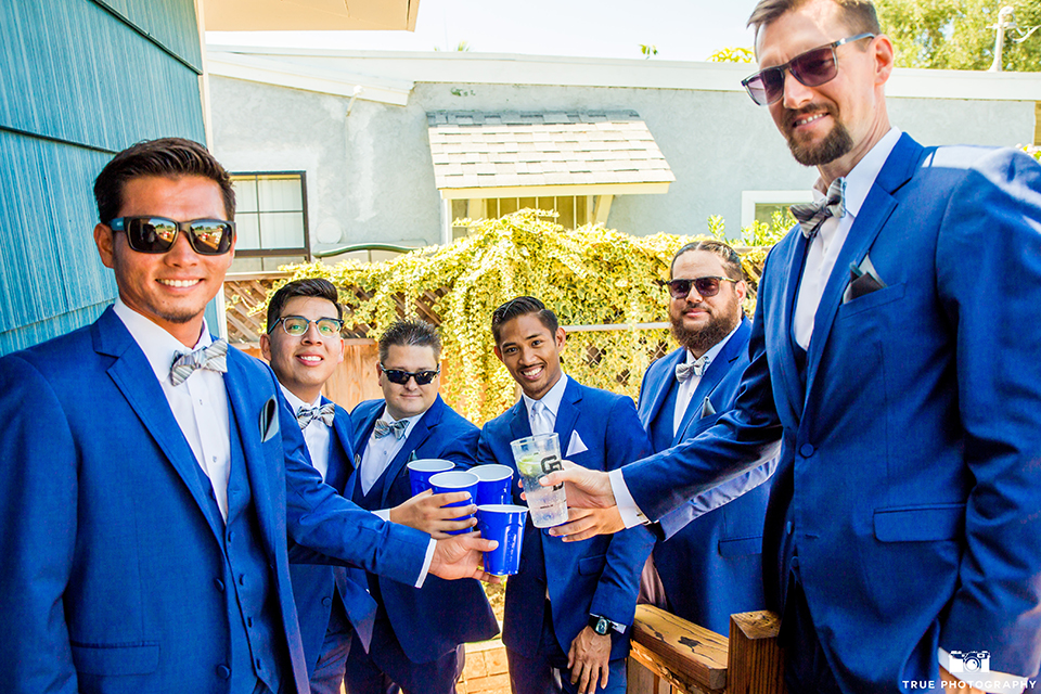 San diego outdoor wedding shoot at bali hai groom cobalt blue notch lapel suit with a white dress shirt and white vest with a long white striped tie and pocket square with white floral boutonniere with groomsmen cobalt blue suits with bow ties holding drinks