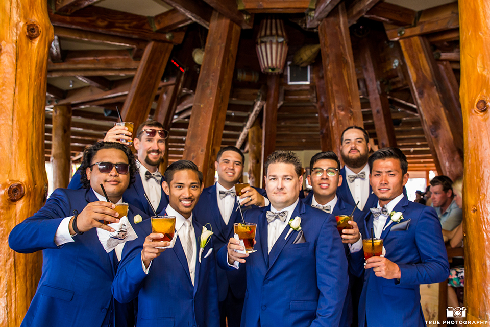 San diego outdoor wedding shoot at bali hai groom cobalt blue notch lapel suit with a white dress shirt and white vest with a long white striped tie and pocket square with white floral boutonniere with groomsmen cobalt blue suits with bow ties holding drinks