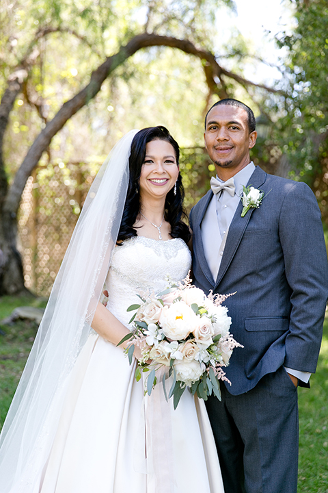 Temecula outdoor wedding at lake oak meadows bride a line strapless gown with lace and detail beading on bodice and long veil with groom grey notch lapel suit with light grey vest and white dress shirt with light grey matching bow tie adn white floral boutonniere standing and hugging bride holding white and green floral bridal bouquet