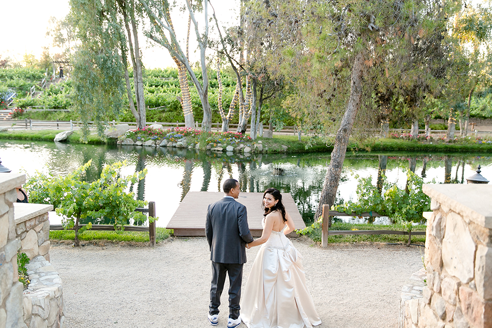 Temecula outdoor wedding at lake oak meadows bride a line strapless gown with lace and detail beading on bodice and long veil with groom grey notch lapel suit with light grey vest and white dress shirt with light grey matching bow tie and white floral boutonniere walking and holding hands bride holding white and green floral bridal bouquet