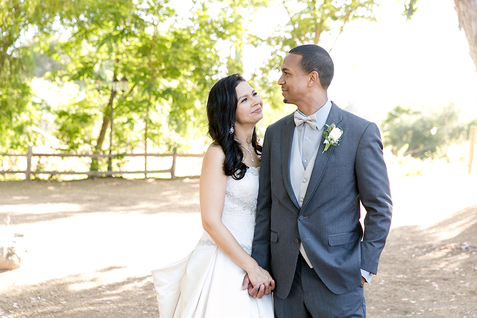 Temecula outdoor wedding at lake oak meadows bride a line strapless gown with lace and detail beading on bodice and long veil with groom grey notch lapel suit with light grey vest and white dress shirt with light grey matching bow tie adn white floral boutonniere standing and holding hands