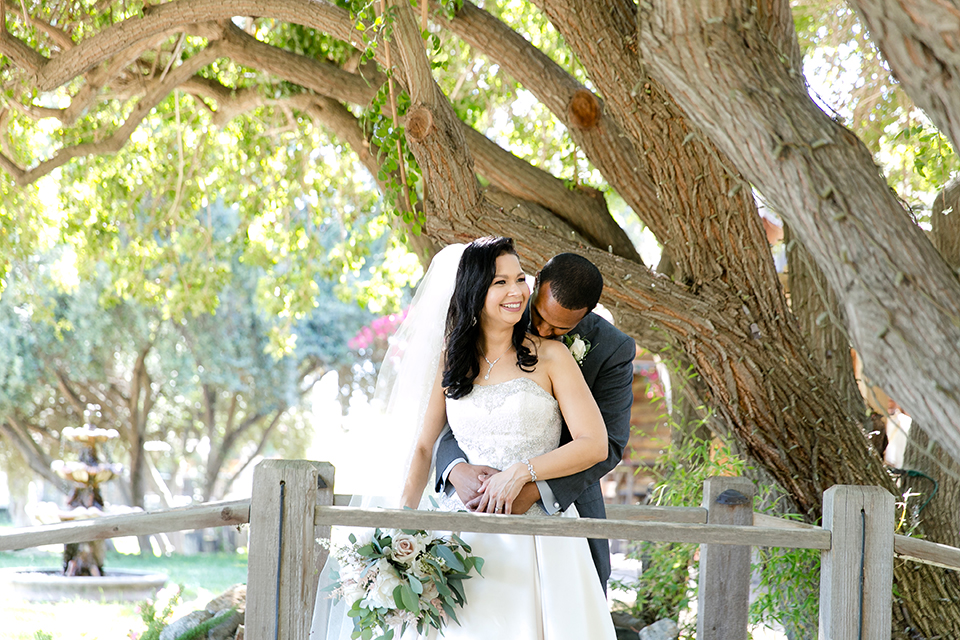 Temecula outdoor wedding at lake oak meadows bride a line strapless gown with lace and detail beading on bodice and long veil with groom grey notch lapel suit with light grey vest and white dress shirt with light grey matching bow tie adn white floral boutonniere standing on bridge and hugging bride holding white and green floral bridal bouquet