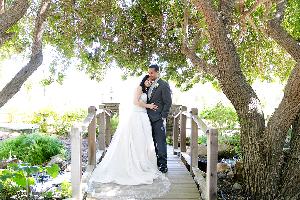 Temecula outdoor wedding at lake oak meadows bride a line strapless gown with lace and detail beading on bodice and long veil with groom grey notch lapel suit with light grey vest and white dress shirt with light grey matching bow tie adn white floral boutonniere standing on bridge and hugging