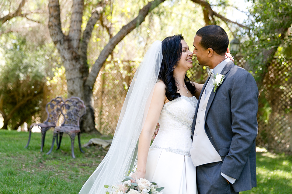 Temecula outdoor wedding at lake oak meadows bride a line strapless gown with lace and detail beading on bodice and long veil with groom grey notch lapel suit with light grey vest and white dress shirt with light grey matching bow tie adn white floral boutonniere standing and hugging bride holding white and green floral bridal bouquet