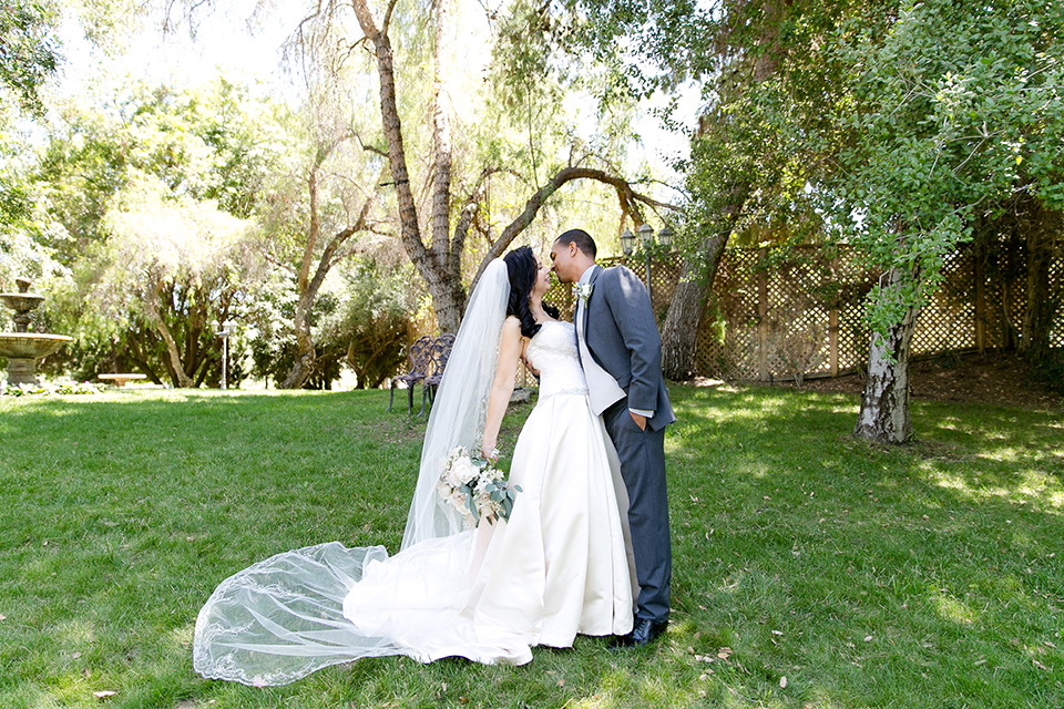 Temecula outdoor wedding at lake oak meadows bride a line strapless gown with lace and detail beading on bodice and long veil with groom grey notch lapel suit with light grey vest and white dress shirt with light grey matching bow tie adn white floral boutonniere standing and hugging bride holding white and green floral bridal bouquet