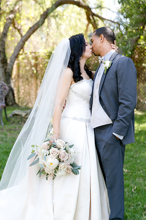 Temecula outdoor wedding at lake oak meadows bride a line strapless gown with lace and detail beading on bodice and long veil with groom grey notch lapel suit with light grey vest and white dress shirt with light grey matching bow tie adn white floral boutonniere standing and kissing bride holding white and green floral bridal bouquet