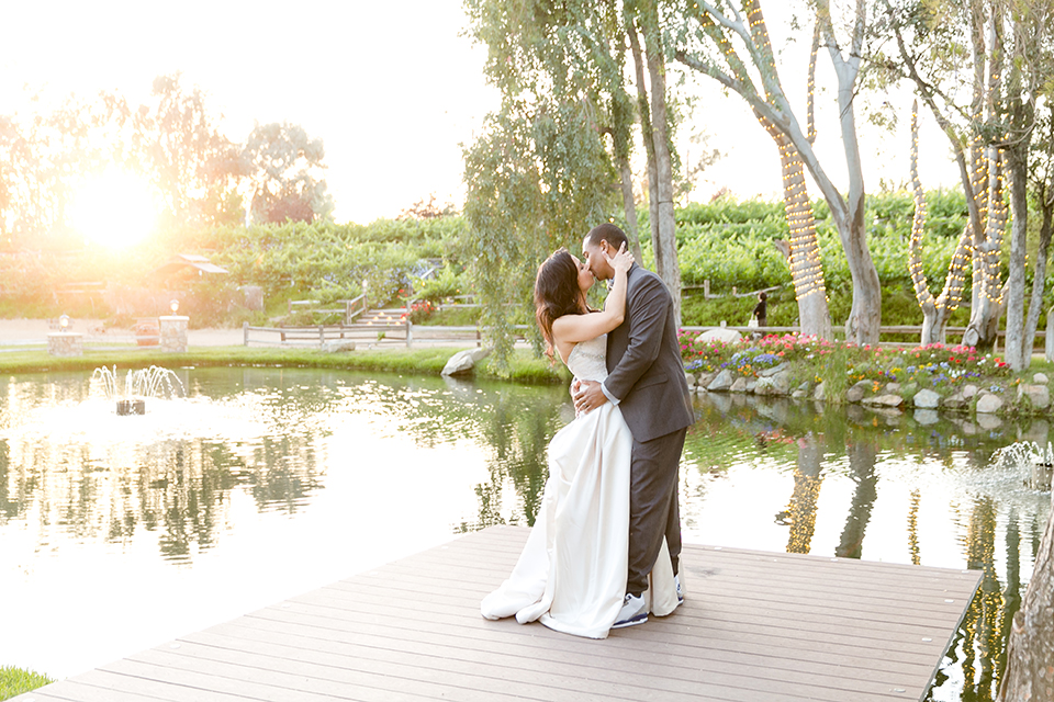 Temecula outdoor wedding at lake oak meadows bride a line strapless gown with lace and detail beading on bodice and long veil with groom grey notch lapel suit with light grey vest and white dress shirt with light grey matching bow tie adn white floral boutonniere standing and kissing