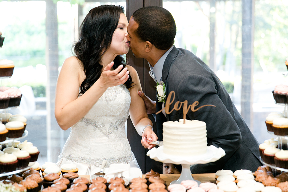 Temecula outdoor wedding at lake oak meadows bride a line strapless gown with lace and detail beading on bodice and long veil with groom grey notch lapel suit with light grey vest and white dress shirt with light grey matching bow tie adn white floral boutonniere bride and groom kissing by wedding cake