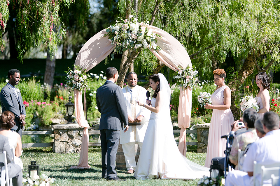 Temecula outdoor wedding at lake oak meadows bride a line strapless gown with lace and detail beading on bodice and long veil with groom grey notch lapel suit with light grey vest and white dress shirt with light grey matching bow tie and white floral boutonniere exchanging vows during ceremony