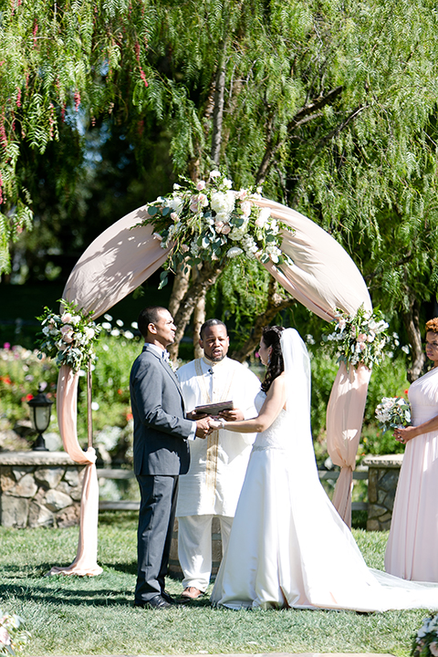 Temecula outdoor wedding at lake oak meadows bride a line strapless gown with lace and detail beading on bodice and long veil with groom grey notch lapel suit with light grey vest and white dress shirt with light grey matching bow tie adn white floral boutonniere holding hands during ceremony