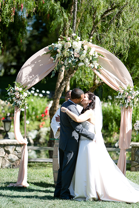 Temecula outdoor wedding at lake oak meadows bride a line strapless gown with lace and detail beading on bodice and long veil with groom grey notch lapel suit with light grey vest and white dress shirt with light grey matching bow tie adn white floral boutonniere standing and kissing during ceremony