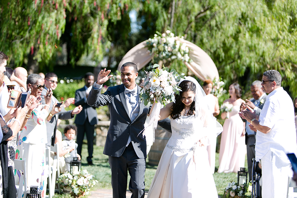 Temecula outdoor wedding at lake oak meadows bride a line strapless gown with lace and detail beading on bodice and long veil with groom grey notch lapel suit with light grey vest and white dress shirt with light grey matching bow tie adn white floral boutonniere walking down the aisle and cheering after ceremony bride holding white and green floral bridal bouquet