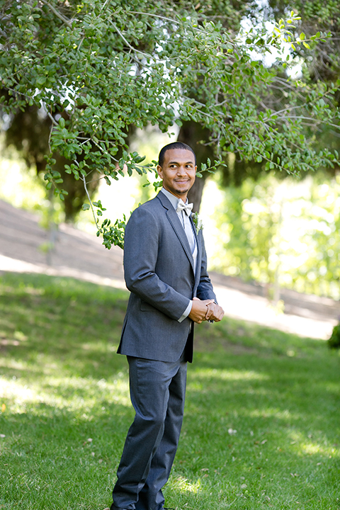 Temecula outdoor wedding at lake oak meadows groom grey notch lapel suit with light grey vest and light grey matching bow tie with white dress shirt and white floral boutonniere standing and smiling