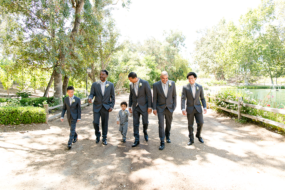 Temecula outdoor wedding at lake oak meadows groom grey notch lapel suit with light grey vest and light grey matching bow tie with white dress shirt and white floral boutonniere walking with groomsmen grey notch lapel suits with light grey vests and ties and ring bearer light grey suit