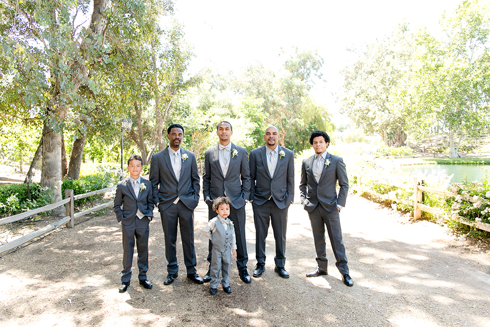 Temecula outdoor wedding at lake oak meadows groom grey notch lapel suit with light grey vest and light grey matching bow tie with white dress shirt and white floral boutonniere standing with groomsmen grey suits with light grey vests and ties and ring bearer light grey suit