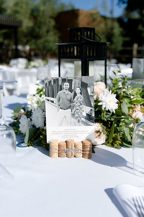 Temecula outdoor wedding at lake oak meadows table set up white table linen with white chairs and white and green flower centerpiece decor with wine cork centerpiece decor with white table numbers and candle lanterns with pictures of bride and groom 