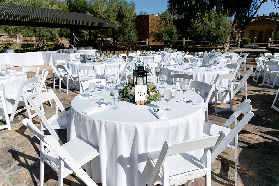 Temecula outdoor wedding at lake oak meadows table set up white table linen with white chairs and white and green flower centerpiece decor with wine cork centerpiece decor with white table numbers and candle lanterns with pictures of bride and groom 