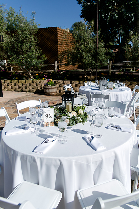 Temecula outdoor wedding at lake oak meadows table set up white table linen with white chairs and white and green flower centerpiece decor with wine cork centerpiece decor with white table numbers and candle lanterns with pictures of bride and groom 