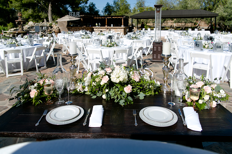 Temecula outdoor wedding at lake oak meadows table set up white table linen with white chairs and white and green flower centerpiece decor with wine cork centerpiece decor with white table numbers and candle lanterns with pictures of bride and groom 