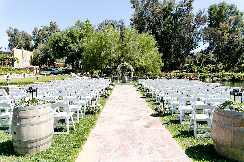 Temecula outdoor wedding at lake oak meadows ceremony set up with white chairs on grass with lanterns along aisle and white and green flower decor with floral arch and wine barrels for decor wedding photo idea for ceremony set up