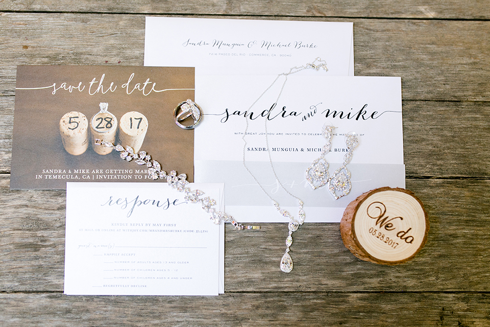 Temecula outdoor wedding at lake oak meadows wedding invitations white with black calligraphy writing and tree stump rustic decor with save the dates and bride jewelry decor on light brown wood background wedding photo idea for invitations