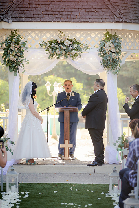 Orange county outdoor summer wedding at the heritage museum bride ball gown with lace straps and a sweetheart neckline with a short tulle skirt and long veil with groom black shawl lapel tuxedo with a white dress shirt and white vest with a white tie and white floral boutonniere standing during ceremony