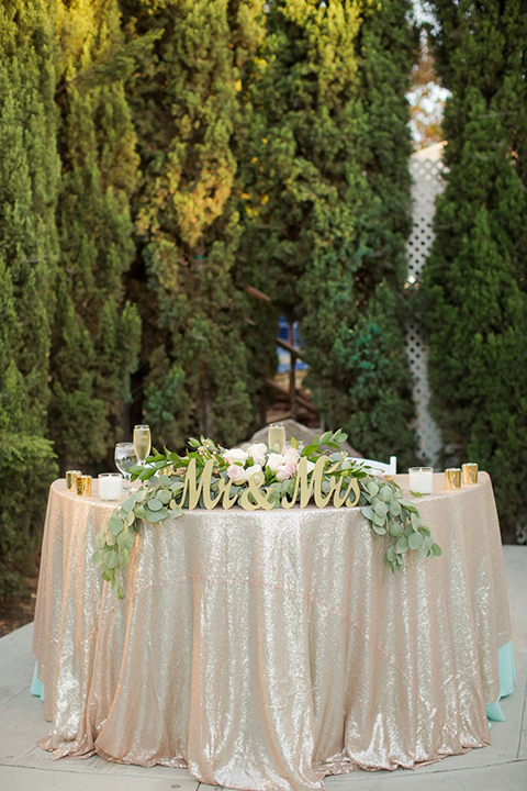 Orange county outdoor summer wedding at the heritage museum wedding reception sweetheart table with champagne sequined table linen and greenery floral decor with white place settings and glassware with gold mr and mrs signs