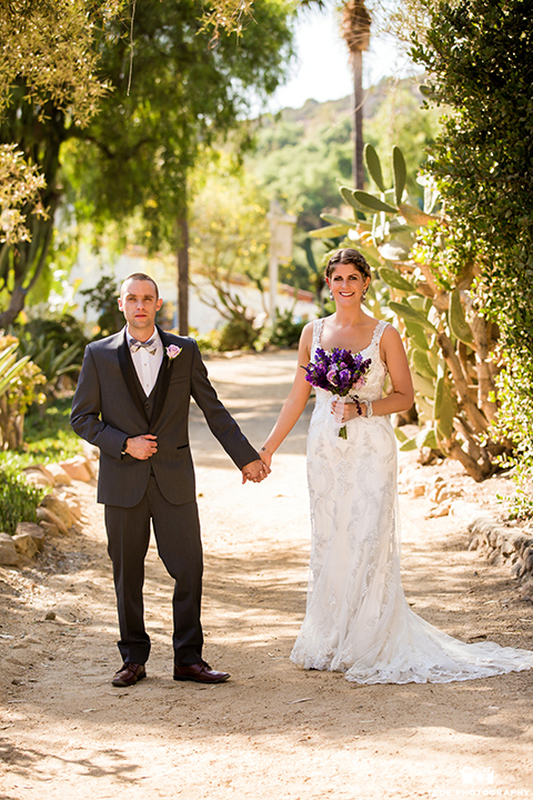 San diego outdoor wedding at leo carillo ranch bride form fitting gown with a sweetheart neckline with thin straps and beaded detail on bodice with groom charcoal grey tuxedo with a black shawl and matching vest with a white dress shirt and plaid bow tie with a white floral boutonniere holding hands and bride holding purple floral bridal bouquet