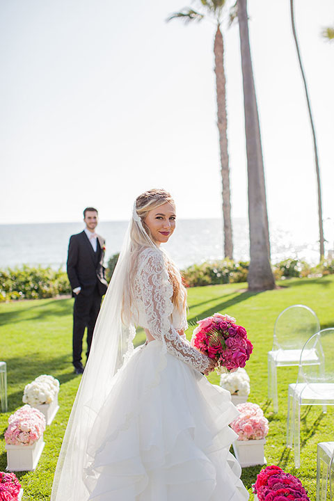 San clemente beach wedding at ole hanson beach club bride two piece wedding gown with lace long sleeves and open back design with a ruffled skirt and long veil with groom navy notch lapel suit with a matching vest and white dress shirt with a blush pink bow tie and white and pink floral boutonniere bride walking down the aisle holding pink floral bridal bouquet