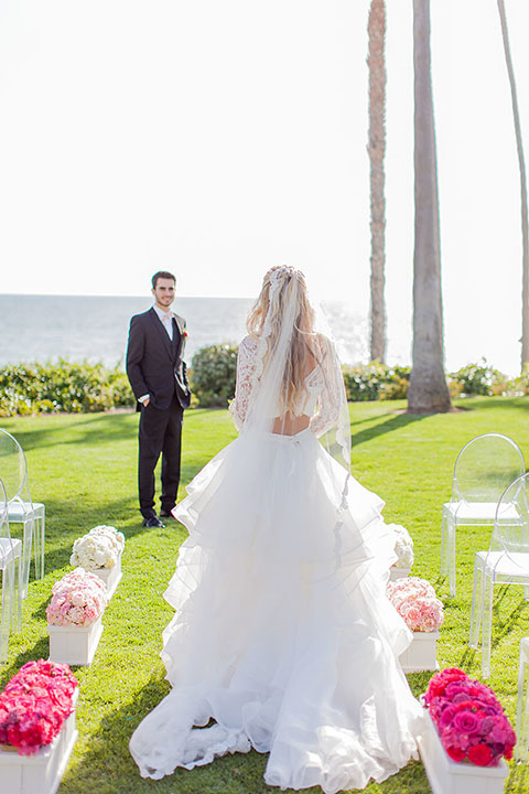 San clemente beach wedding at ole hanson beach club bride two piece wedding gown with lace long sleeves and open back design with a ruffled skirt and long veil with groom navy notch lapel suit with a matching vest and white dress shirt with a blush pink bow tie and white and pink floral boutonniere bride walking down the aisle during ceremony
