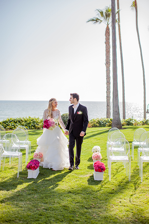 San clemente beach wedding at ole hanson beach club bride two piece wedding gown with lace long sleeves and open back design with a ruffled skirt and long veil with groom navy notch lapel suit with a matching vest and white dress shirt with a blush pink bow tie and white and pink floral boutonniere holding hands and walking down the aisle after ceremony