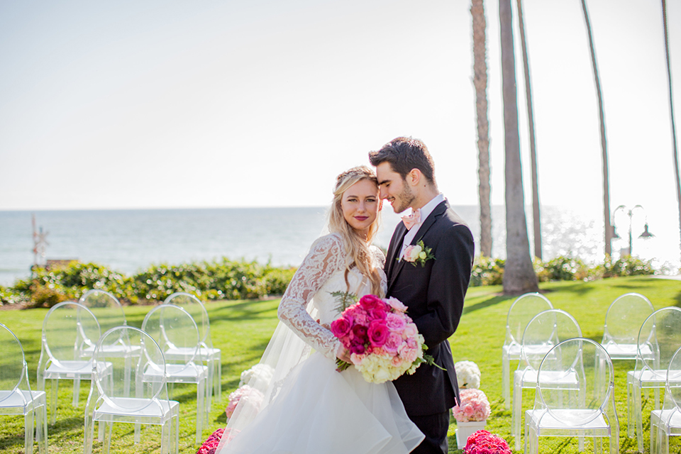 San clemente beach wedding at ole hanson beach club bride two piece wedding gown with lace long sleeves and open back design with a ruffled skirt and long veil with groom navy notch lapel suit with a matching vest and white dress shirt with a blush pink bow tie and white and pink floral boutonniere hugging in aisle after ceremony bride holding pink floral bridal bouquet