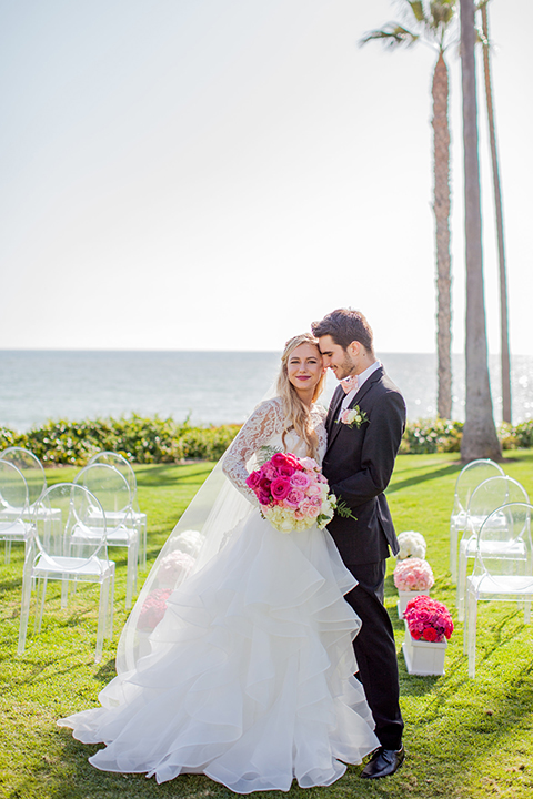 San clemente beach wedding at ole hanson beach club bride two piece wedding gown with lace long sleeves and open back design with a ruffled skirt and long veil with groom navy notch lapel suit with a matching vest and white dress shirt with a blush pink bow tie and white and pink floral boutonniere hugging in aisle after ceremony bride holding pink floral bridal bouquet