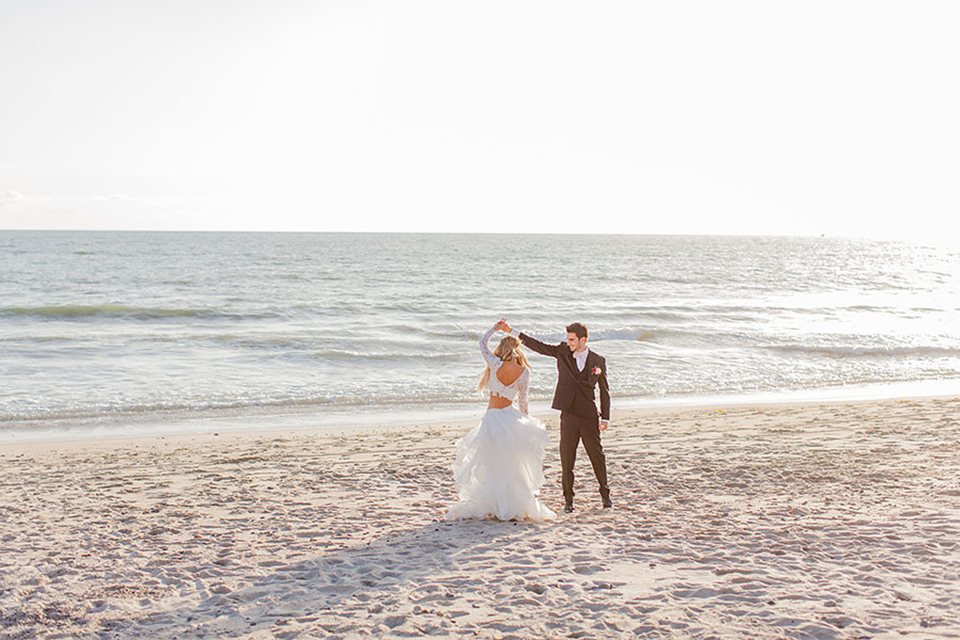San clemente beach wedding at ole hanson beach club bride two piece wedding gown with lace long sleeves and open back design with a ruffled skirt and long veil with groom navy notch lapel suit with a matching vest and white dress shirt with a blush pink bow tie and white and pink floral boutonniere dancing on the beach