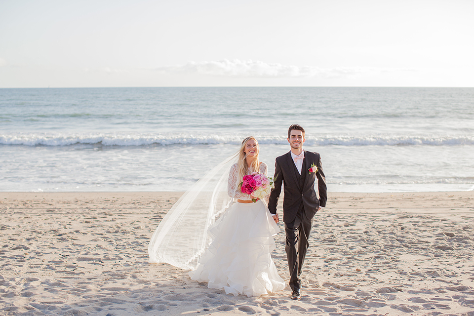 San clemente beach wedding at ole hanson beach club bride two piece wedding gown with lace long sleeves and open back design with a ruffled skirt and long veil with groom navy notch lapel suit with a matching vest and white dress shirt with a blush pink bow tie and white and pink floral boutonniere walking and holding hands on the beach and bride holding pink floral bridal bouquet