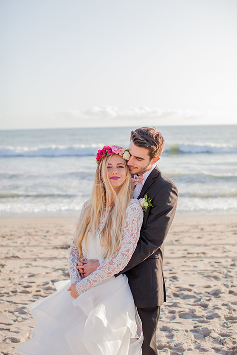 San clemente beach wedding at ole hanson beach club bride two piece wedding gown with lace long sleeves and open back design with a ruffled skirt and long veil with groom navy notch lapel suit with a matching vest and white dress shirt with a blush pink bow tie and white and pink floral boutonniere hugging on the beach bride wearing pink floral crown