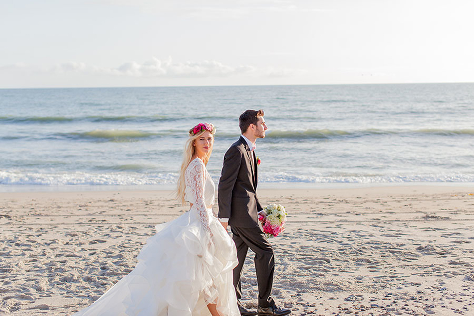 San clemente beach wedding at ole hanson beach club bride two piece wedding gown with lace long sleeves and open back design with a ruffled skirt and long veil with groom navy notch lapel suit with a matching vest and white dress shirt with a blush pink bow tie and white and pink floral boutonniere walking on the beach and holding hands and groom holding pink floral bridal bouquet