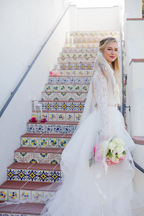 San clemente beach wedding at ole hanson beach club bride two piece wedding gown with lace long sleeves and open back design with a ruffled skirt and long veil and white and pink floral bridal bouquet standing on stairs