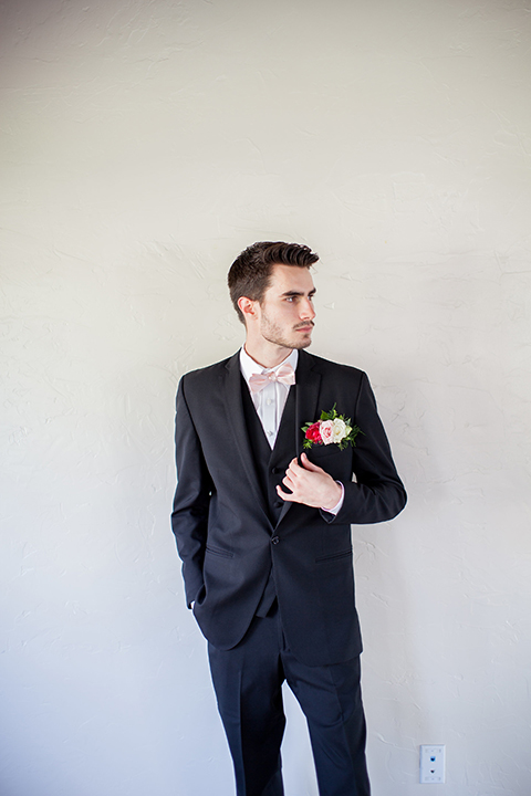 San clemente beach wedding shoot at ole hanson beach club groom navy notch lapel suit with a matching vest and white dress shirt with a blush pink bow tie and pink and white floral boutonniere standing with hands in pockets