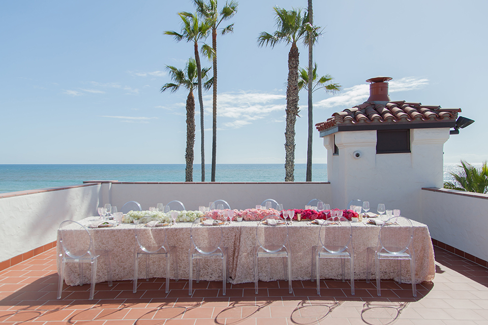 San clemente beach wedding shoot at ole hanson beach club table set up with white table floral linen and blush pink flower centerpiece decor with white and gold place settings with pink menus and glassware with clear ghost chairs