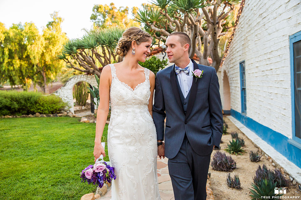 San diego outdoor wedding at leo carillo ranch bride form fitting gown with a sweetheart neckline with thin straps and beaded detail on bodice with groom charcoal grey tuxedo with a black shawl and matching vest with a white dress shirt and plaid bow tie with a white floral boutonniere holding hands and bride holding purple floral bridal bouquet