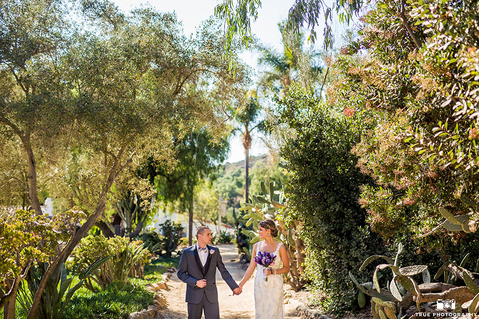 San diego outdoor wedding at leo carillo ranch bride form fitting gown with a sweetheart neckline with thin straps and beaded detail on bodice with groom charcoal grey tuxedo with a black shawl and matching vest with a white dress shirt and plaid bow tie with a white floral boutonniere holding hands and bride holding purple floral bridal bouquet