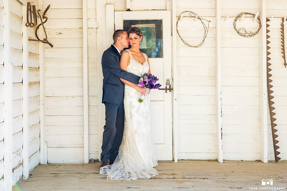 San diego outdoor wedding at leo carillo ranch bride form fitting gown with a sweetheart neckline with thin straps and beaded detail on bodice with groom charcoal grey tuxedo with a black shawl and matching vest with a white dress shirt and plaid bow tie with a white floral boutonniere hugging and bride holding purple floral bridal bouquet