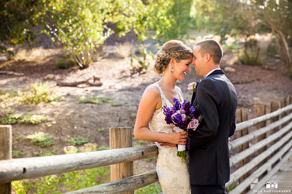 San diego outdoor wedding at leo carillo ranch bride form fitting gown with a sweetheart neckline with thin straps and beaded detail on bodice with groom charcoal grey tuxedo with a black shawl and matching vest with a white dress shirt and plaid bow tie with a white floral boutonniere hugging and bride holding purple floral bridal bouquet