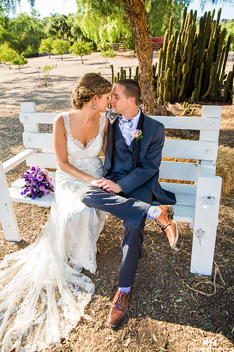 San diego outdoor wedding at leo carillo ranch bride form fitting gown with a sweetheart neckline with thin straps and beaded detail on bodice with groom charcoal grey tuxedo with a black shawl and matching vest with a white dress shirt and plaid bow tie with a white floral boutonniere sitting on white bench with purple floral bridal bouquet