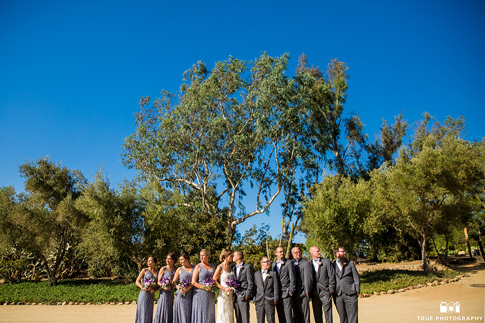 San diego outdoor wedding at leo carillo ranch bride form fitting gown with a sweetheart neckline with thin straps and beaded detail on bodice with groom charcoal grey tuxedo with a black shawl and matching vest with a white dress shirt and plaid bow tie with a white floral boutonniere with wedding party bridesmaids long lavender dresses with beading detail and groomsmen charcoal grey tuxedos with bow ties