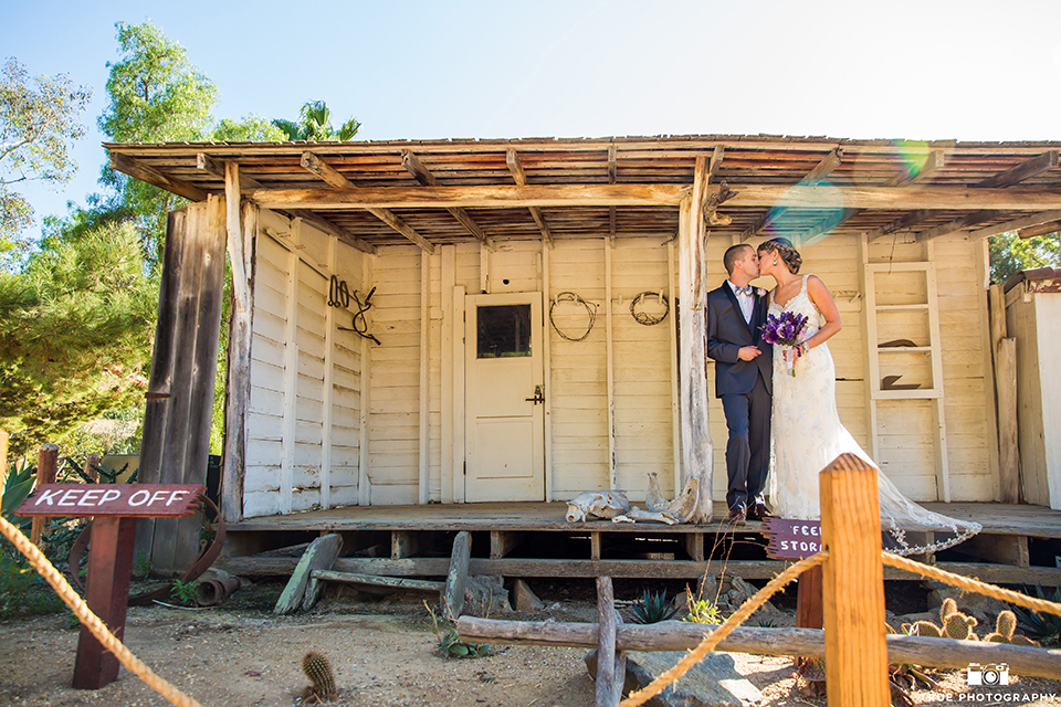 San diego outdoor wedding at leo carillo ranch bride form fitting gown with a sweetheart neckline with thin straps and beaded detail on bodice with groom charcoal grey tuxedo with a black shawl and matching vest with a white dress shirt and plaid bow tie with a white floral boutonniere hugging bride holding purple floral bridal bouquet