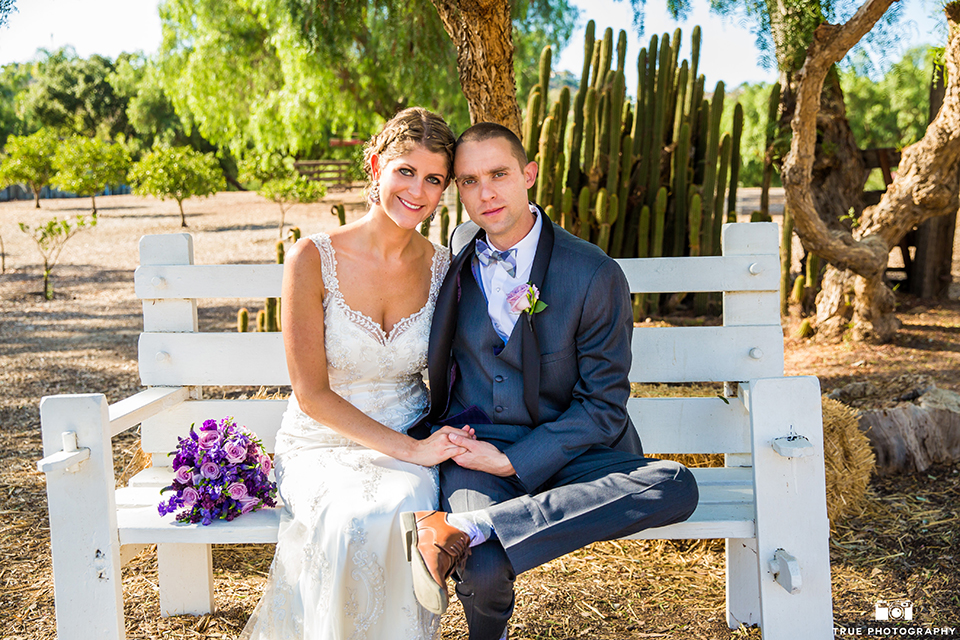 San diego outdoor wedding at leo carillo ranch bride form fitting gown with a sweetheart neckline with thin straps and beaded detail on bodice with groom charcoal grey tuxedo with a black shawl and matching vest with a white dress shirt and plaid bow tie with a white floral boutonniere sitting on bench with purple floral bridal bouquet