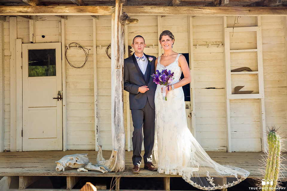 San diego outdoor wedding at leo carillo ranch bride form fitting gown with a sweetheart neckline with thin straps and beaded detail on bodice with groom charcoal grey tuxedo with a black shawl and matching vest with a white dress shirt and plaid bow tie with a white floral boutonniere hugging bride holding purple floral bridal bouquet