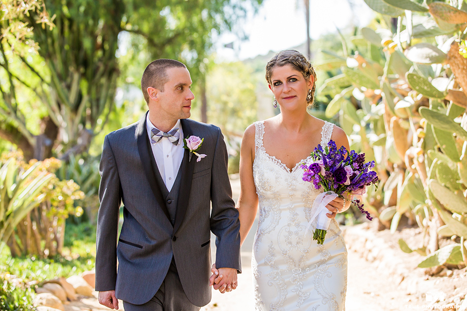 San diego outdoor wedding at leo carillo ranch bride form fitting gown with a sweetheart neckline with thin straps and beaded detail on bodice with groom charcoal grey tuxedo with a black shawl and matching vest with a white dress shirt and plaid bow tie with a white floral boutonniere holding hands and bride holding purple floral bridal bouquet